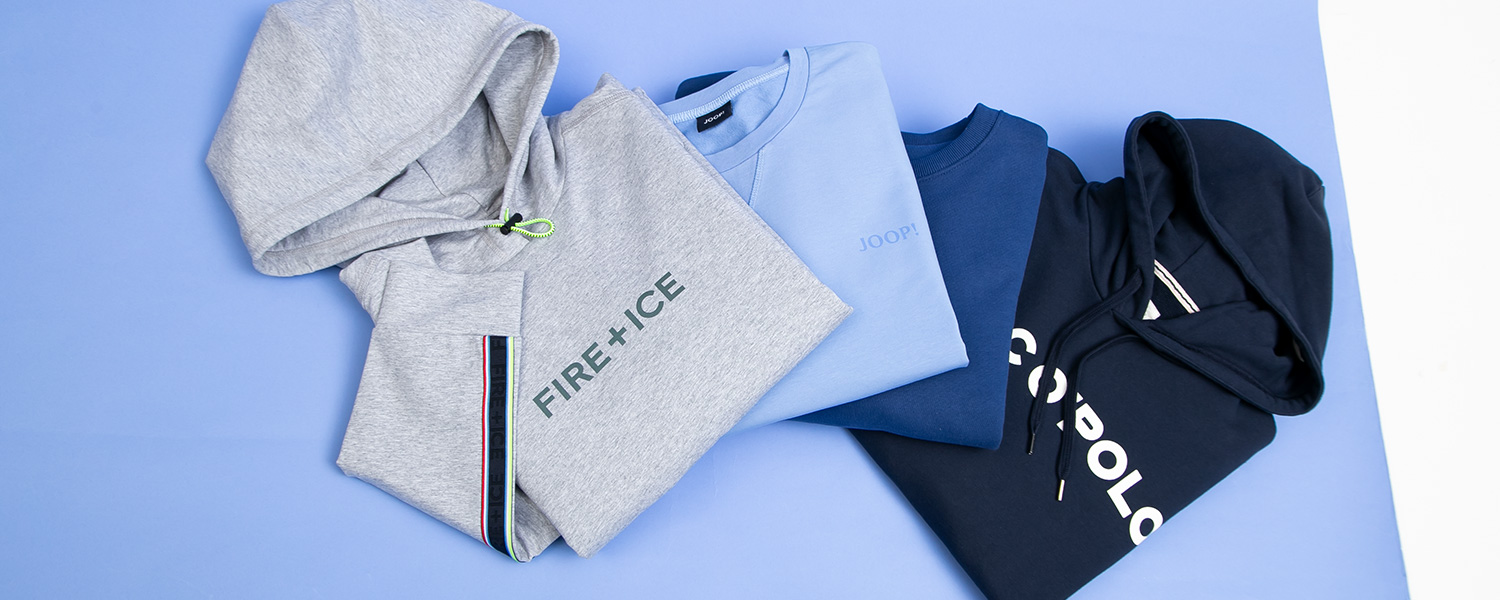 Auf dem Bild steht ein Mann vor alten Eisenbahnwaggons und trägt eine graue Sweatjacke unter einem Mantel.