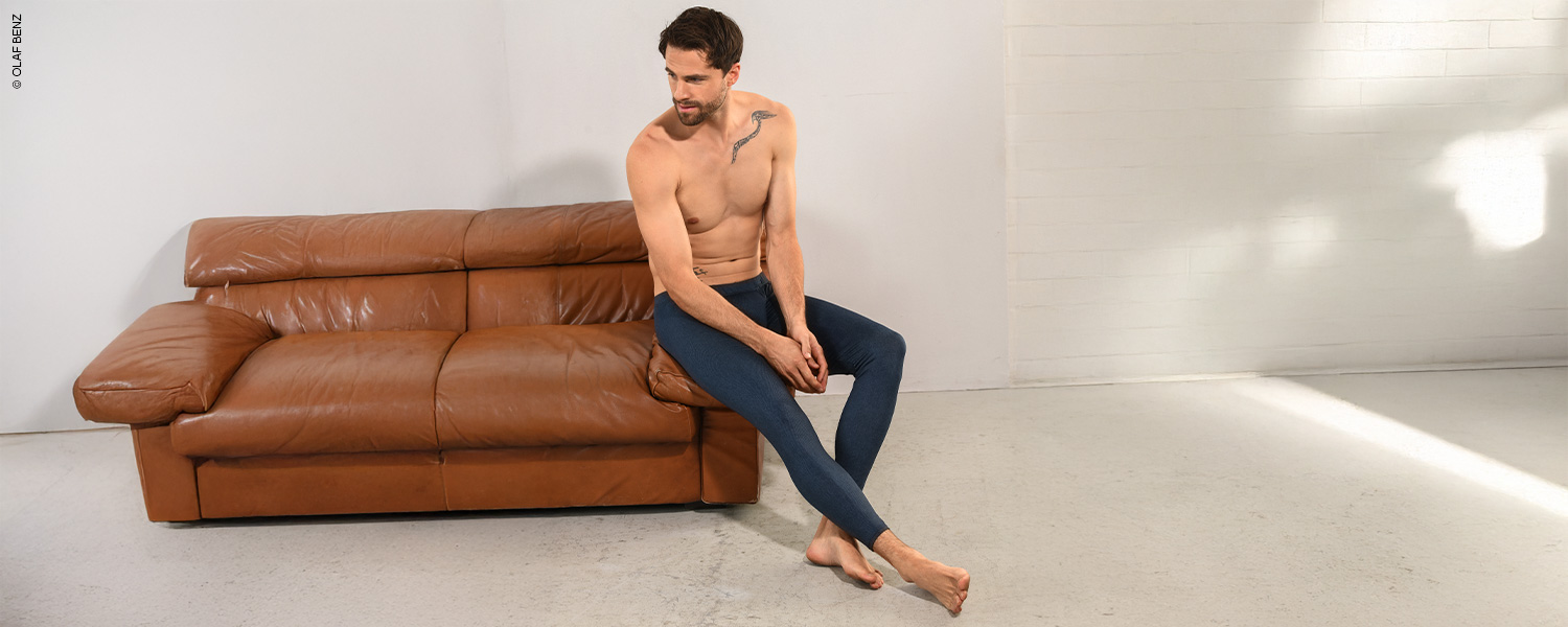 Ein Mann in dunkler langer Unterhose und langem Shirt sitzt neben einer Frau in heller Unterwäsche auf dem Balkon.