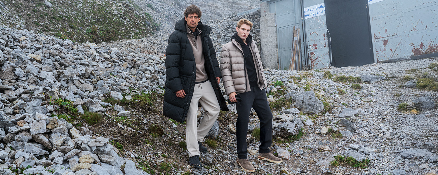 Zwei Männer in gepolsterten Daunenjacken, schwarz und beige, auf steinigem Hang