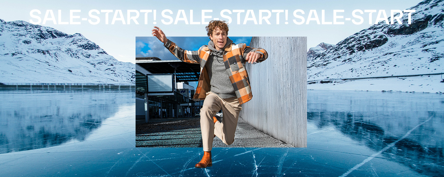 Karierte Wolljacke in Grau und Orange, lässig getragen. Im Hintergrund eine winterliche Berglandschaft zu gefrorenem See