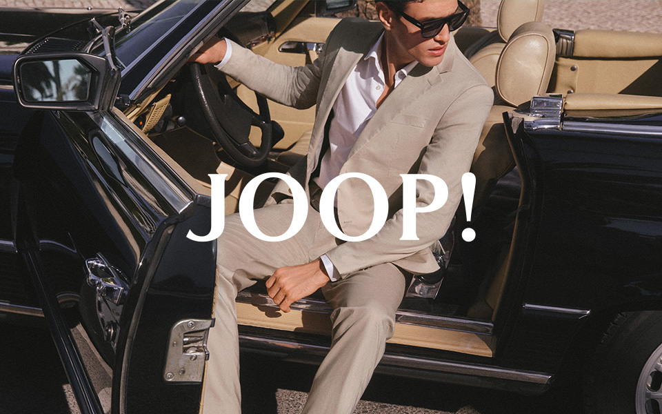 Mann im beigen Anzug mit weißem Hemd und Mokassins, lässig im Auto sitzend