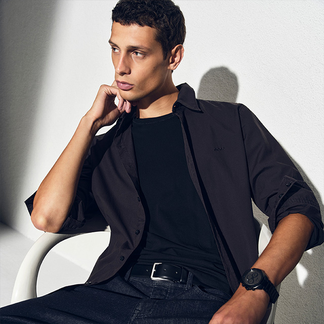 Mann in schwarzem Freizeithemd aus Baumwolle, lässig offen getragen über schwarzem T-Shirt, sitzt auf Stuhl