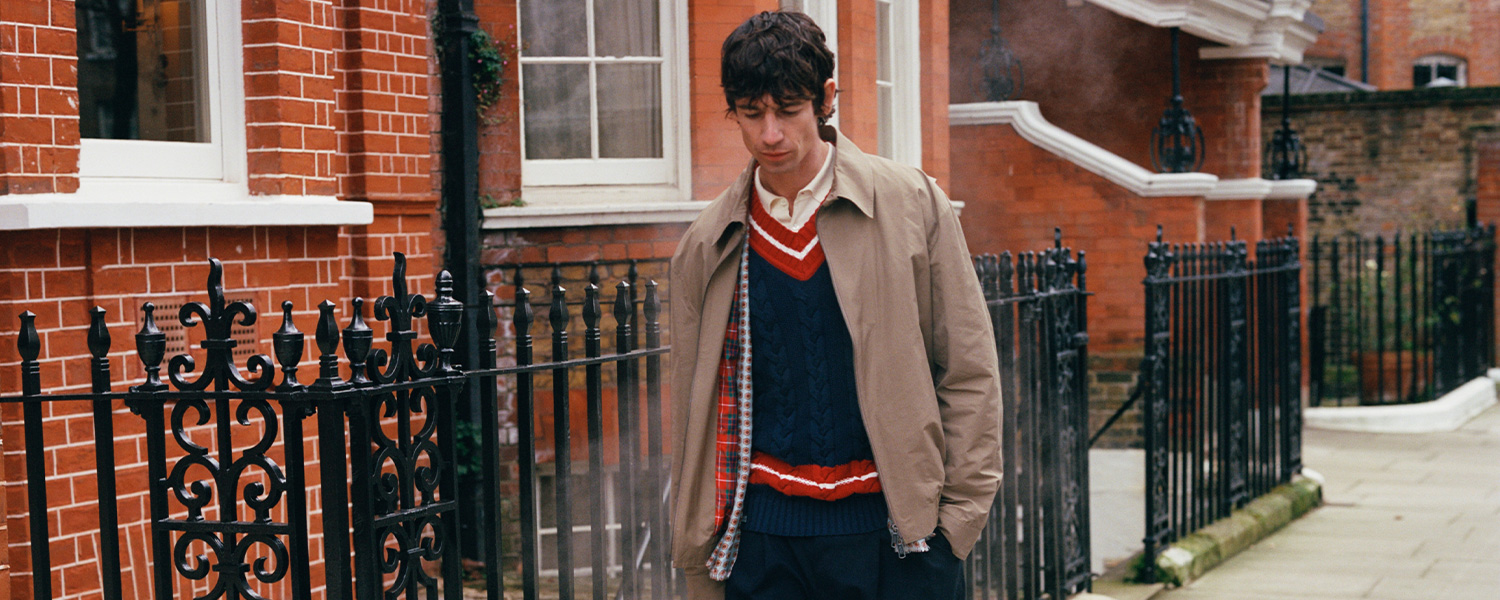 Beiger Trenchcoat über blauem Strickpullover mit V-Ausschnitt und rotem Streifen, lässig getragen