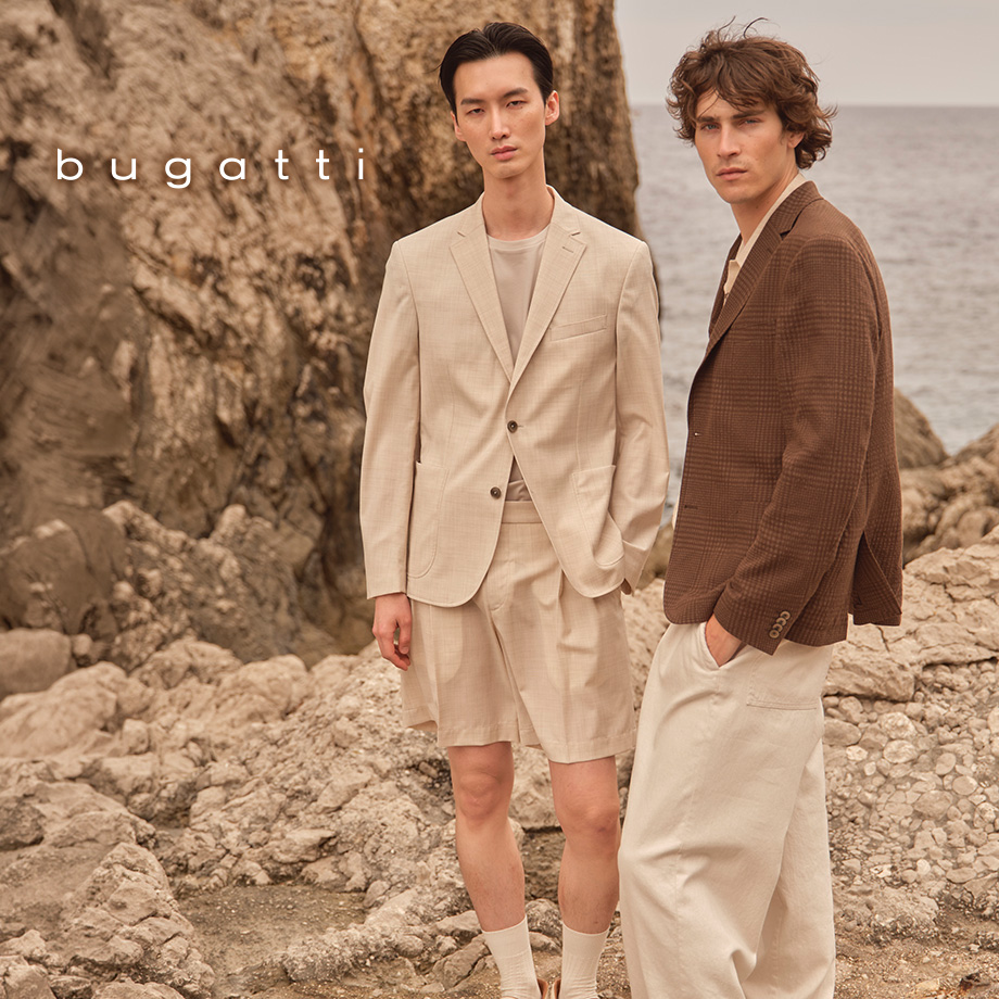 Zwei Männer am Felsenstrand in beigefarbenem Anzug mit Shorts und braunem Sakko über T-Shirt