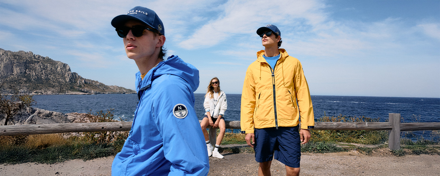 Zwei Männer in blauer und gelber Windjacke mit Basecaps, am Meer entlang spazierend.