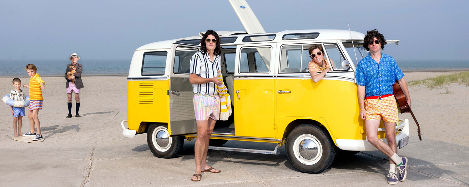 Sommer-Outfit: Mann in gestreifter Badeshorts und schwarz-weiß gestreiftem Hemd vor gelbem VW-Bus am Strand