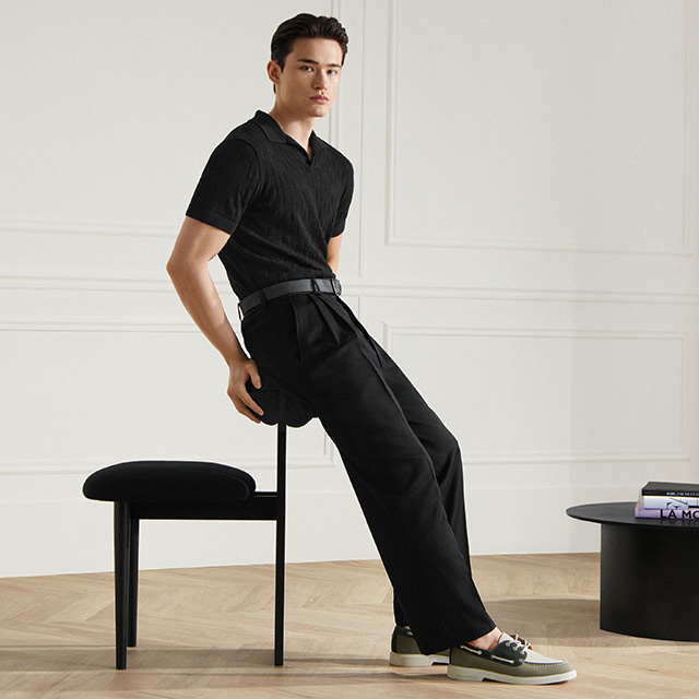 Mann in schwarzem Polohemd und Bundfaltenhose, leger auf Stuhl sitzend im eleganten Raum