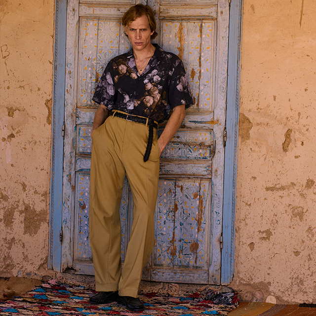 Mann in gemustertem, kurzärmligem Hemd und beiger Stoffhose, lässig angelehnt
