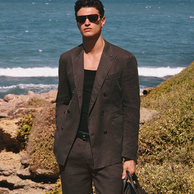 Mann im dunkelbraunen Leinenblazer mit schwarzem Shirt am Strand