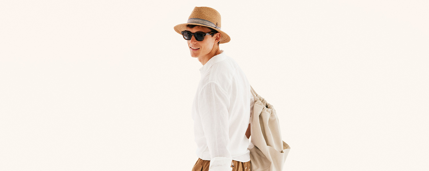 Mann in weißem Leinenhemd, Strohhut und Sonnenbrille, mit beiger Tasche