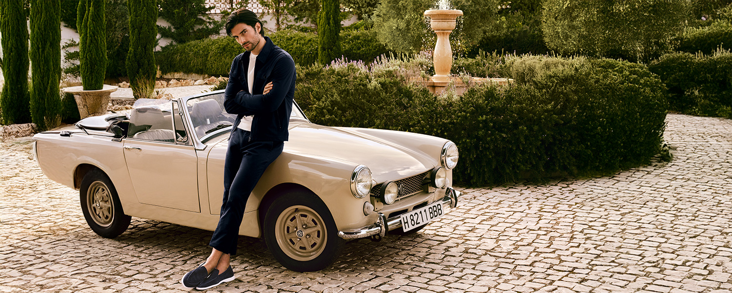 Mann in dunkelblauer Jacke und Hose, lässig an cremefarbenem Oldtimer im Garten lehnend