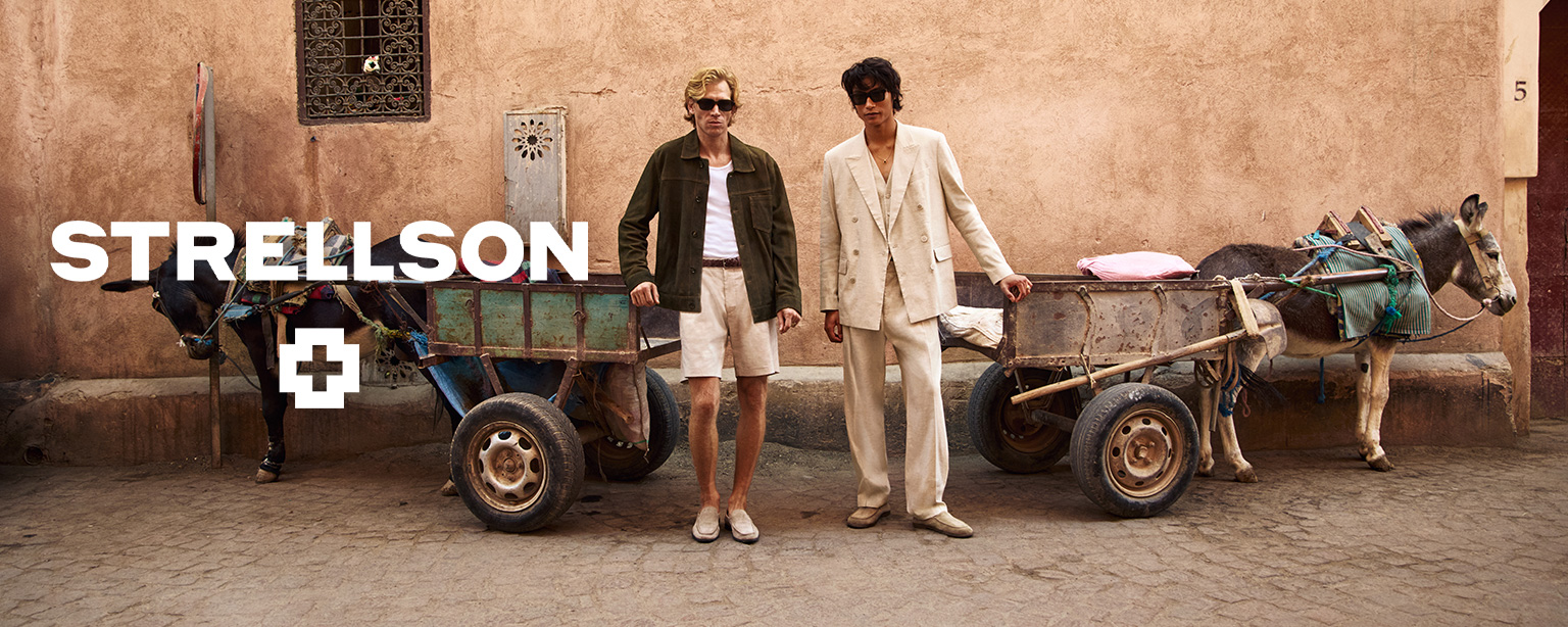 Herr in beigem Leinenanzug mit Sonnenbrille, leger getragen, neben Mann in olivgrüner Jacke und Shorts