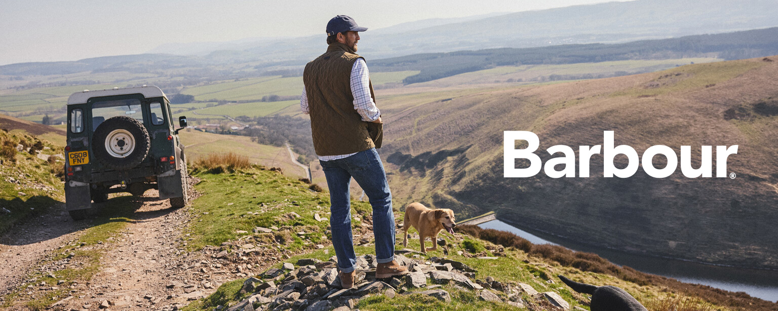 Mann in Outdoor-Weste und Jeans, blickt in die Landschaft, neben Geländewagen und Hund