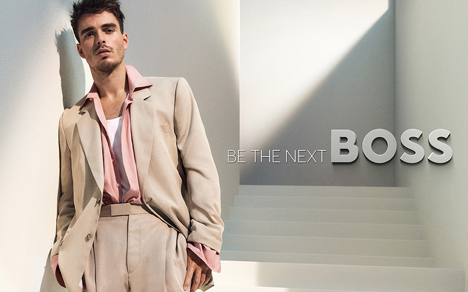 Mann in hellbeigem Anzug und rosa Hemd, lässig stehend in hellem Raum.