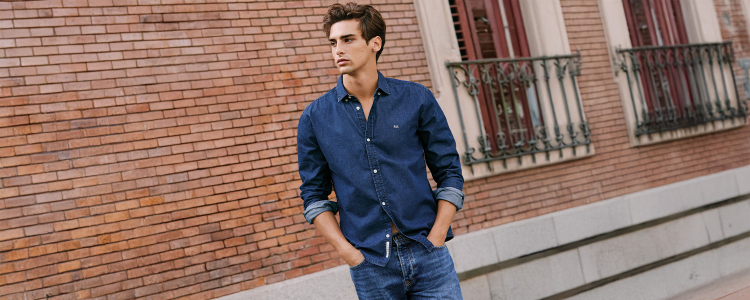 Mann in dunkelblauem Jeanshemd, lässig mit umgeschlagenen Ärmeln und Jeanshose vor Backsteinwand