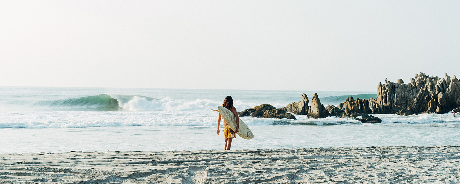 Surfer in gelber Badeshorts mit Surfbrett am Strand vor felsiger Kulisse