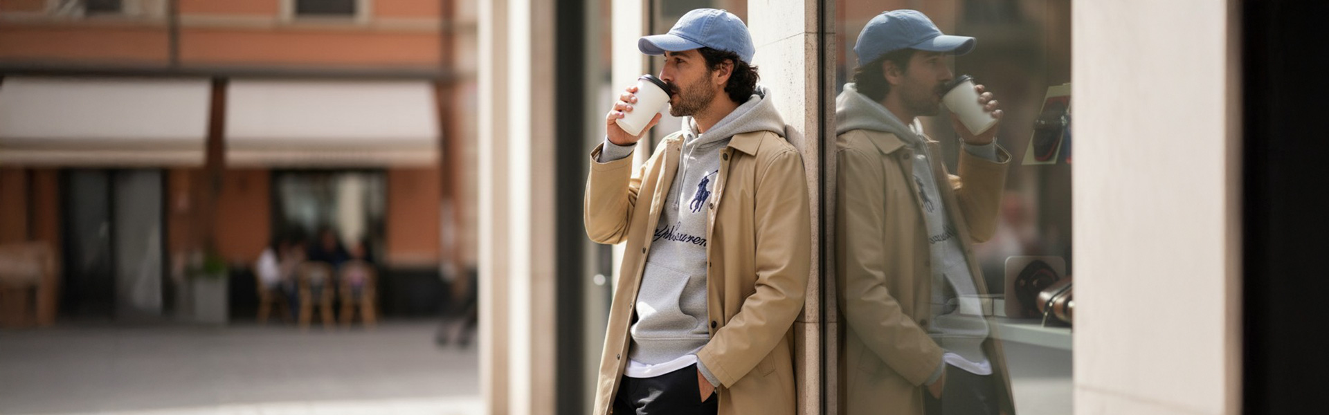 Mann im beigen Mantel und grauem Hoodie, lässig stehend, trinkt Kaffee