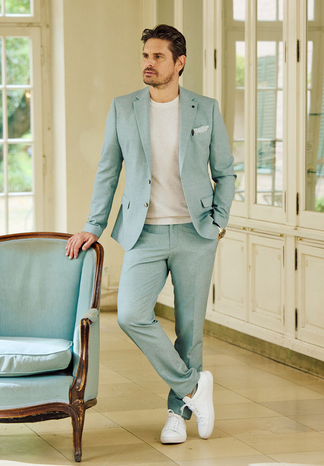 Eleganter Herrenanzug in Hellblau mit weißem Pullover und Sneakers getragen, lässig im Raum präsentiert