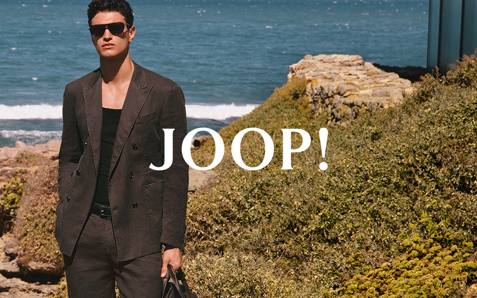 Mann im braunen Leinenanzug mit Sonnenbrille am Strand, hält schwarze Reisetasche