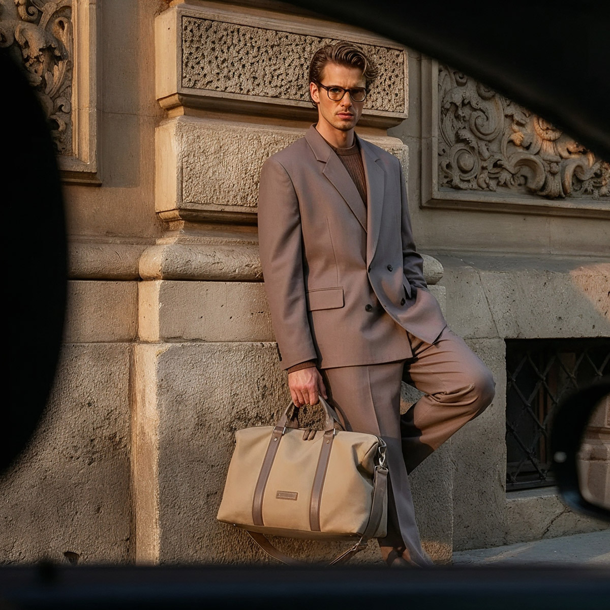 Mann in taupefarbenem Anzug und Brille mit großer Tasche, angelehnt an historische Steinmauer