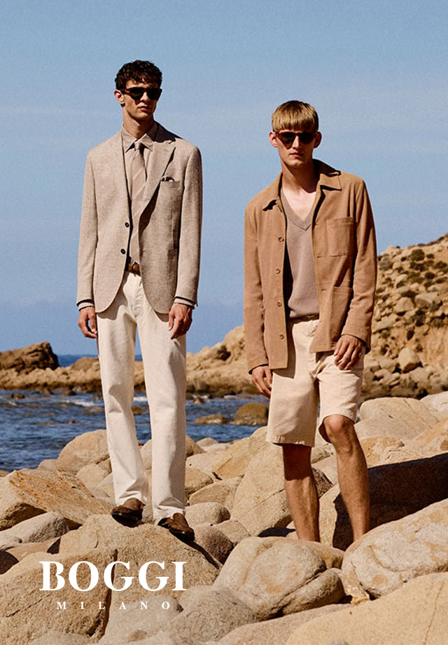 Zwei Männer, einer in beigem Leinen-Blazer mit weißer Hose und Hemd am Strand. Der andere trägt ein braunes Overhirt, dazu beige Shorts und ein dunkelbeiges Shirt. 