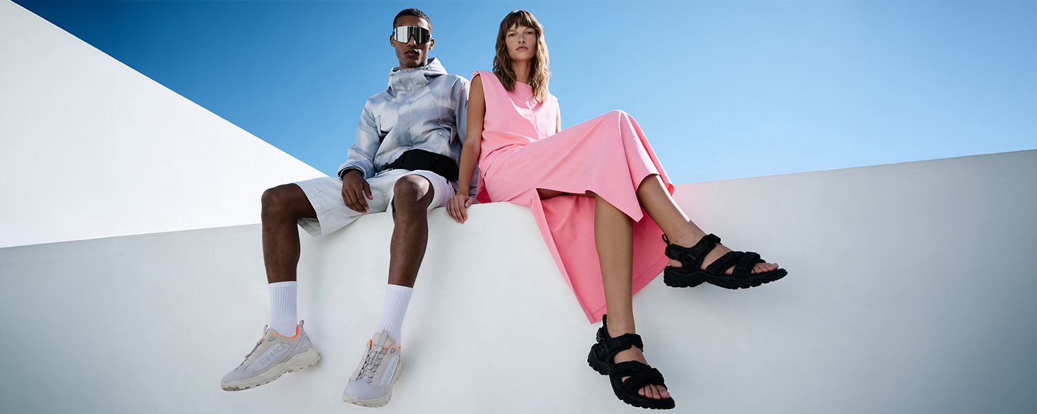 Casual-Look mit grauer Windjacke und weißen Shorts auf Mauer sitzend, ergänzt durch Sneakers.