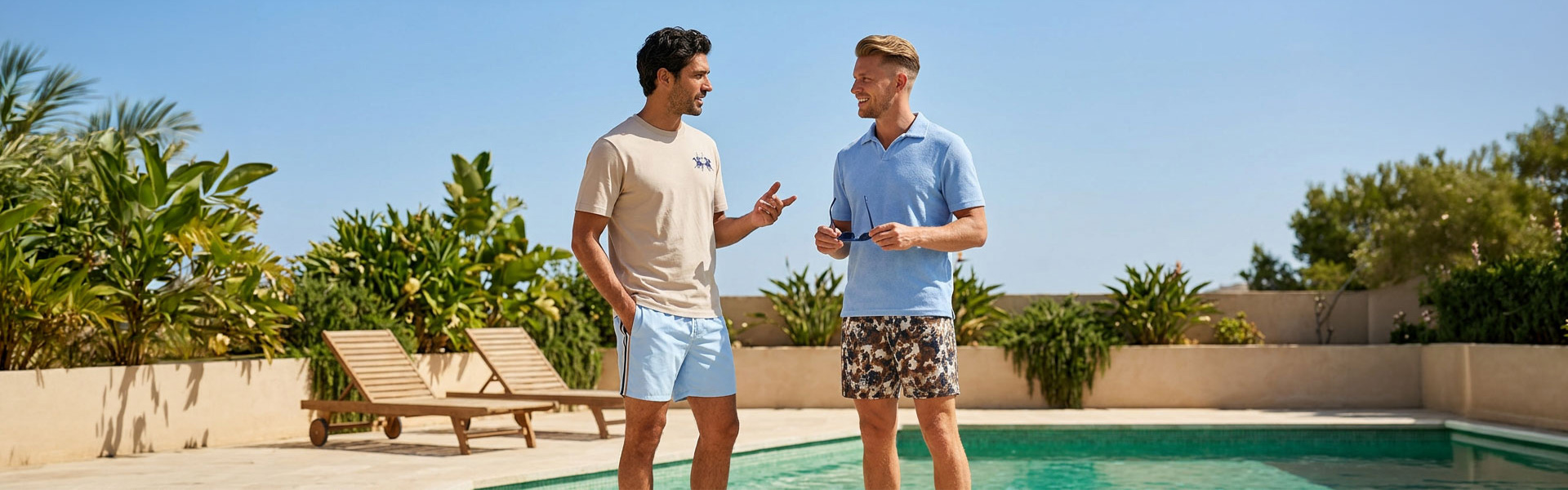 Zwei Männer am Pool, einer trägt hellblaues Poloshirt mit gemusterten Shorts, der andere beigefarbenes T-Shirt mit hellblauer Shorts.