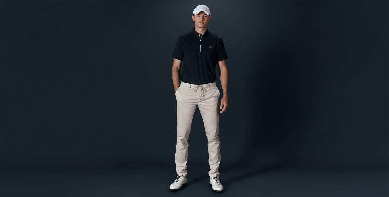 Mann in schwarzem Poloshirt und beigefarbenen Golf Chino vor dunklem Hintergrund