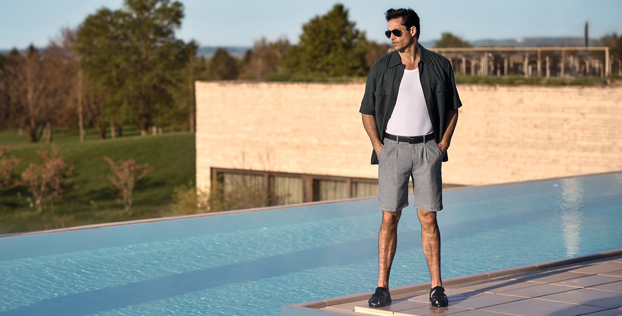 Mann in grauen Alberto Shorts und weißem Tanktop, mit offenem Hemd steht lässig am Pool