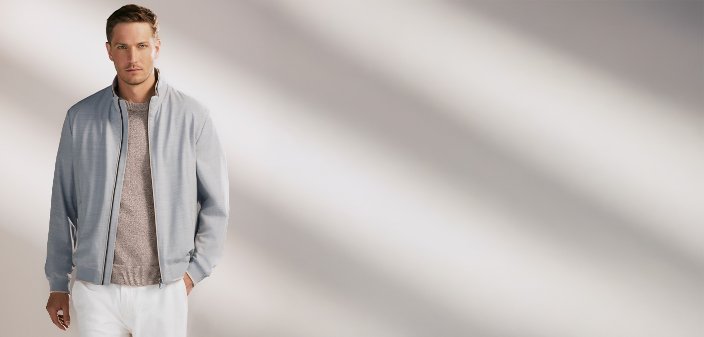 Hellgraue Herrenjacke aus Baumwolle, lässig offen über beigem Pullover, kombiniert mit weißen Hosen