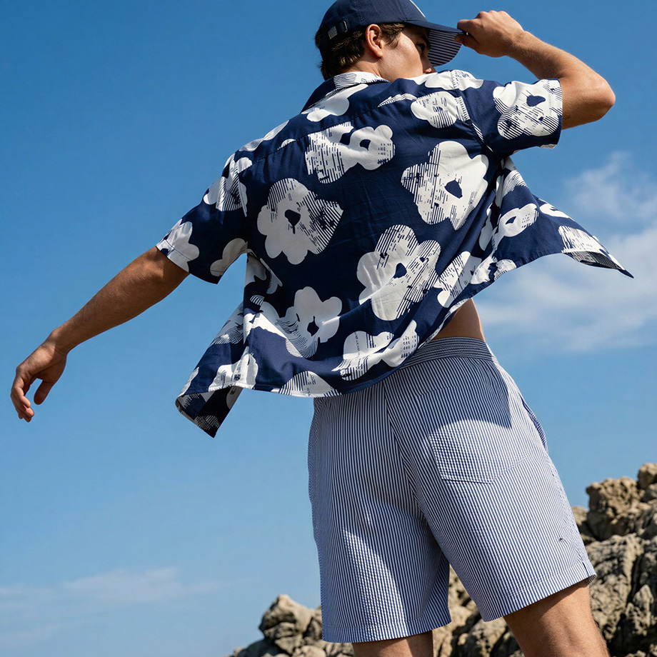 Mann in blauem Hawaiihemd mit weißen Blumen, gestreifte Shorts, sommerlich am Strand