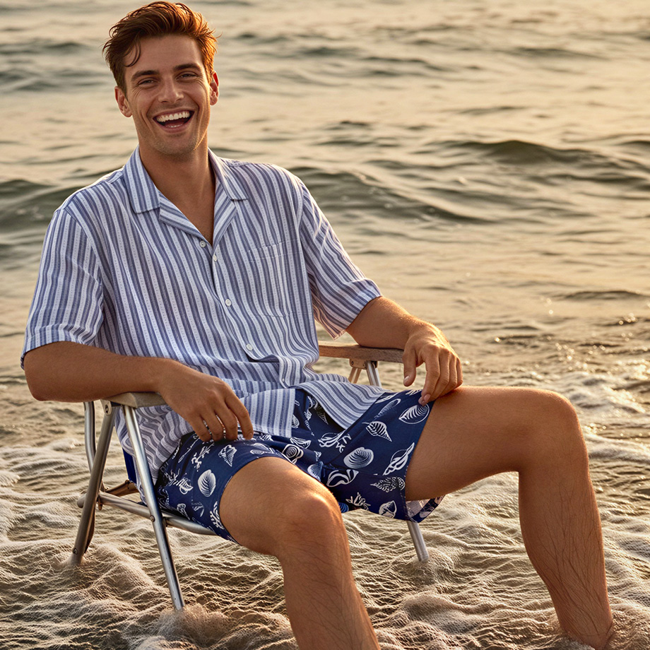 Mann am Strand in hellblau gestreiftem Kurzarmhemd und blauen Badeshorts mit Muschelmuster