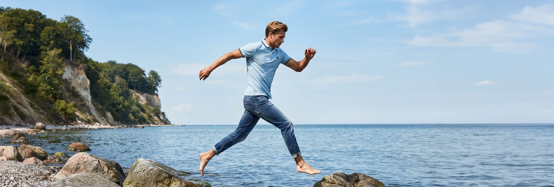 Mann in hellblauem Poloshirt und Jeans springt am Meer über Steine