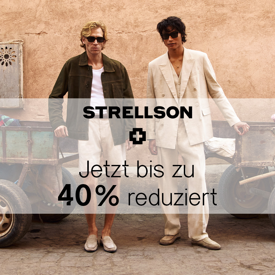 Zwei Männer in beigen Anzügen, einer mit grünem Blazer, lässig vor rustikaler Kulisse