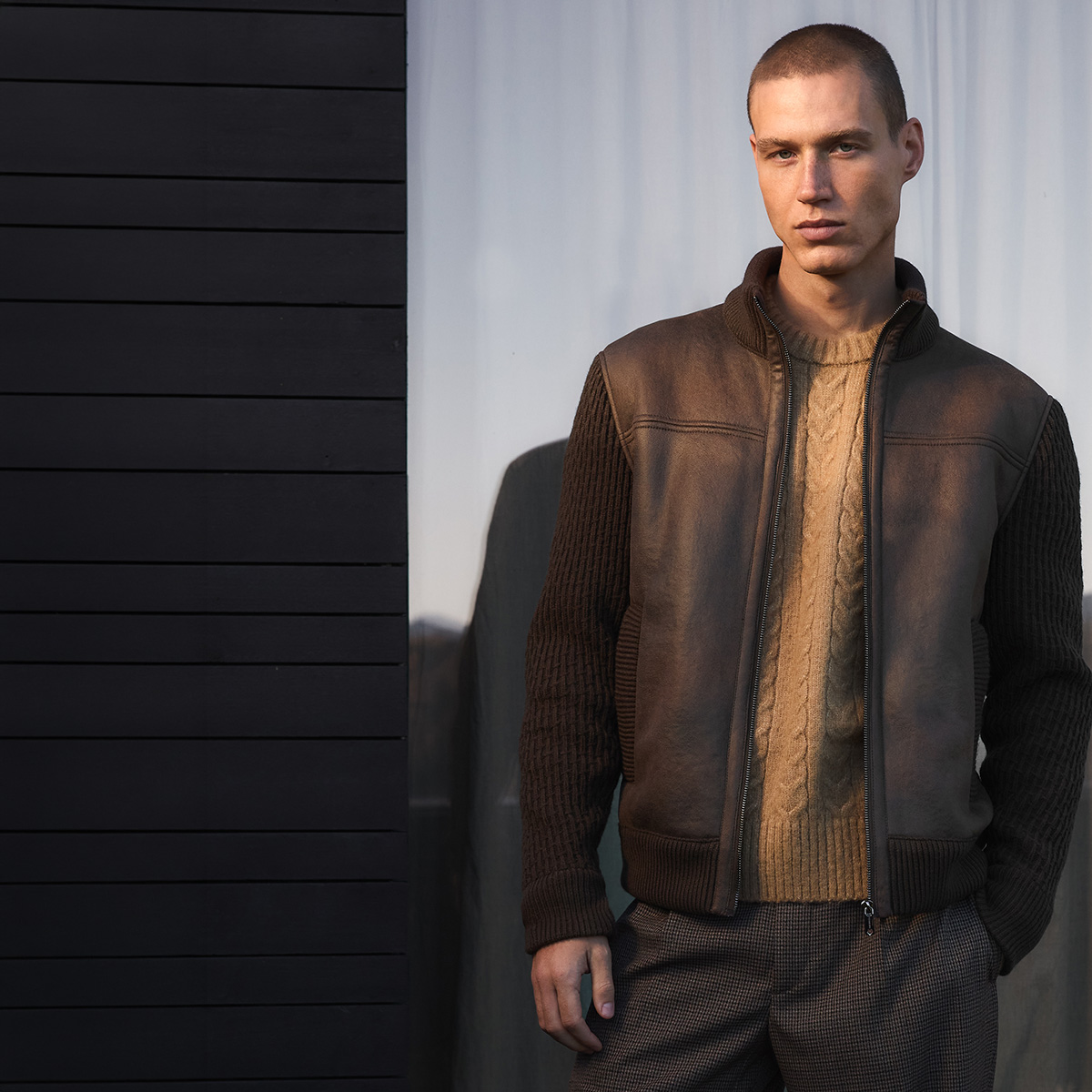 Mann in brauner Lederjacke und Strickpullover, lässig vor modernem Hintergrund