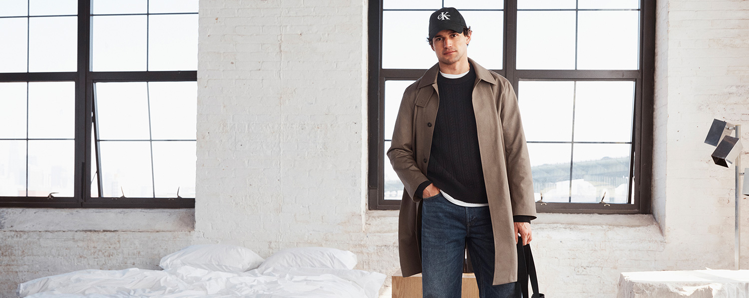 Mann in beigem Trenchcoat mit dunkelblauem Pullover und Jeans, casual getragen vor hellem Fenster.
