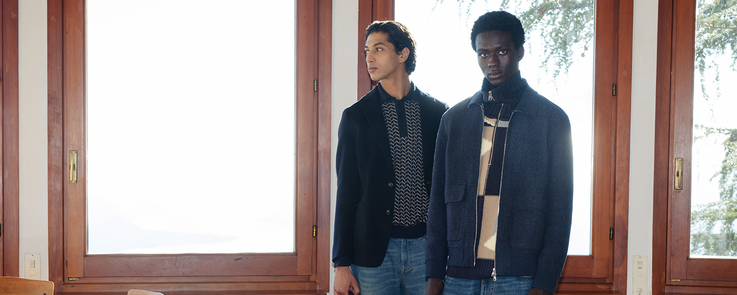 Herren in dunkelblauen Strickpullovern und Jacken vor Fenster stehend, casual elegant.