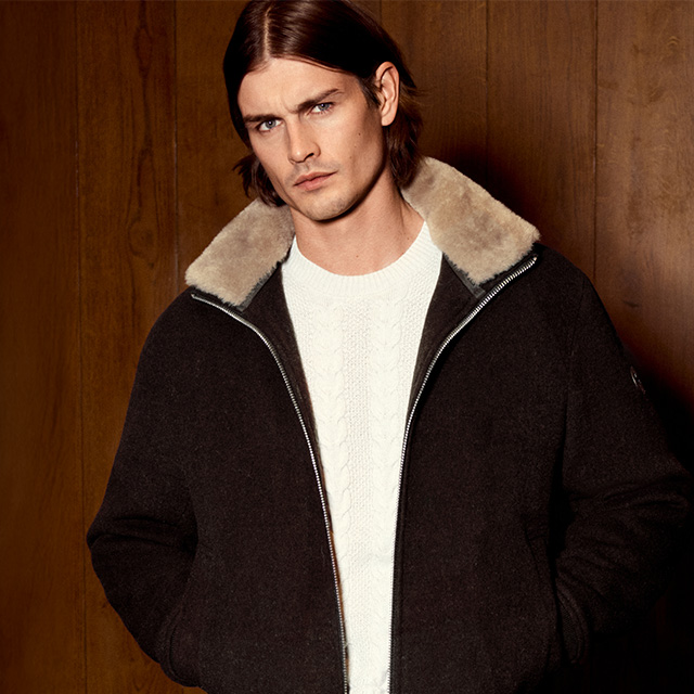 Mann in dunkelbrauner Wolljacke mit Stehkragen und weißem Strickpullover vor Holzpaneelen