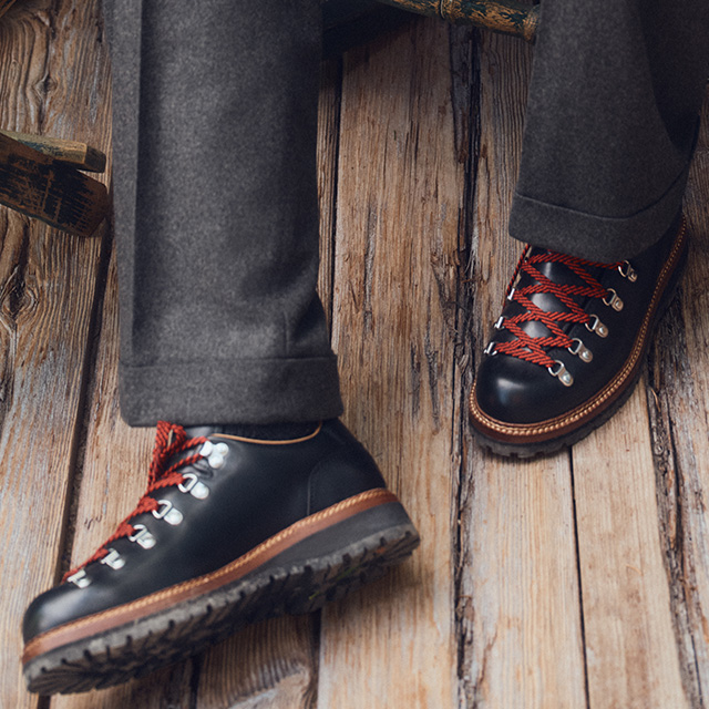 Schwarze Leder-Einsteigerstiefel mit roter Schnürung, getragen mit grauer Wollhose auf Holzuntergrund