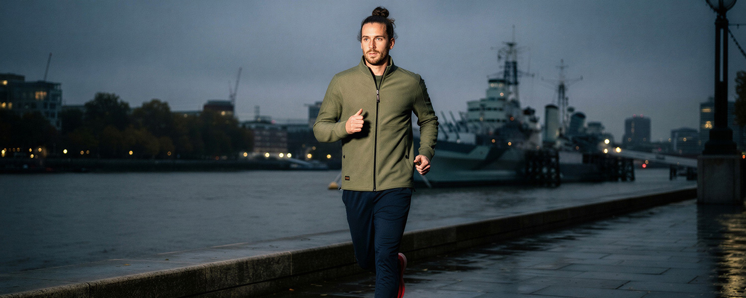Mann in olivgrüner Fleecejacke mit Reißverschluss beim Joggen an der Uferpromenade in der Dämmerung