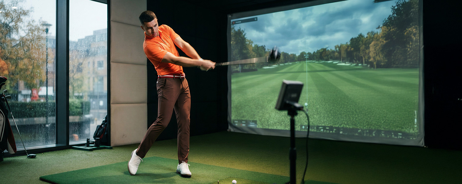 Mann in orangefarbenem Poloshirt und brauner Chino-Hose beim Golfsimulator