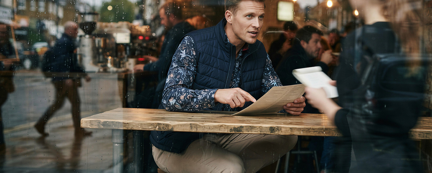 Mann in marineblaue Steppweste und gemusterten Hemd mit Paisley-Print im Café, Smart-Casual-Look