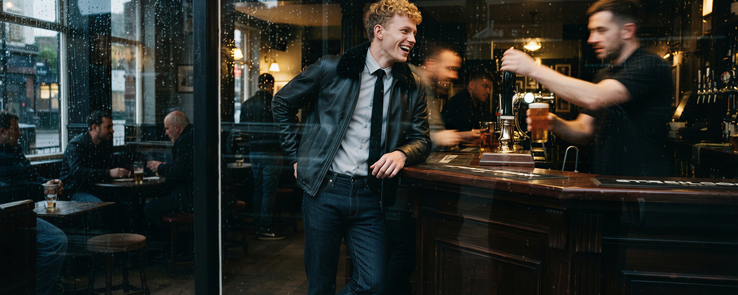 Mann in schwarzer Lederjacke über weißem Hemd mit Krawatte und dunkler Jeans in urbaner Bar
