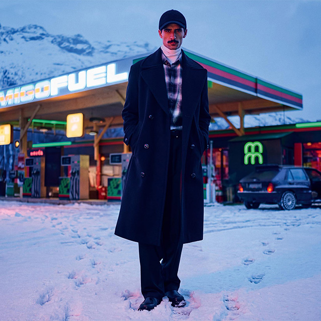 Mann in schwarzem Wollmantel und karierter Jacke vor Tankstelle im Schnee
