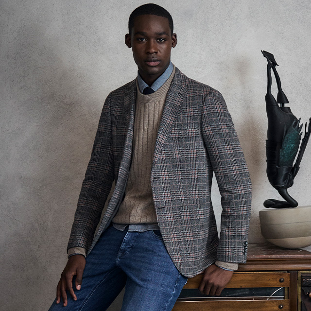 Man in checked blazer with beige pullover and shirt, sitting casually on desk