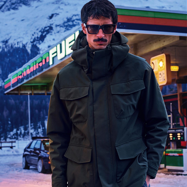 Herr in olivgrüner Outdoor-Jacke, Kapuze und großen Taschen, vor Tankstelle im Winter